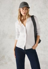 Weißer gerippter Cardigan mit Knöpfen, über dunkelblauen Jeans getragen. Das Model trägt außerdem eine graue Strickmütze und eine schwarze Umhängetasche.