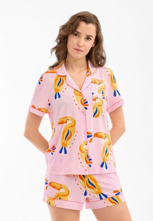 Vrouw draagt een roze kort mouw button-up shirt en shorts set met geel toekan vogelprint, staand tegen een witte achtergrond.