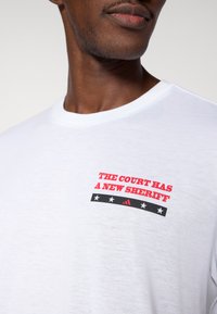 Hvid bomuld T-shirt med rød tekst "THE COURT HAS A NEW SHERIFF" over en sort-hvid grafik med stjerner og et Adidas-logo.