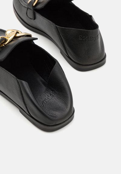 Μαύρες δερμάτινες μοκέτες με τετράγωνες μύτες, με χαρακτηριστική χρυσή αλυσίδα μπροστά και ανάγλυφο λογότυπο στην περιοχή της φτέρνας.