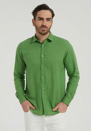 Homme aux cheveux courts et foncés avec une barbe, portant une chemise verte à manches longues boutonnée et un pantalon blanc, debout les mains dans les poches.