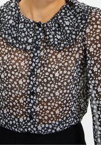 Černá průsvitná blůza s volánkovým límcem, zdobená bílým květinovým vzorem a černými knoflíky na přední straně. Měkká, lehká tkanina.