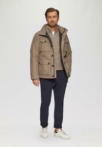 Graubraune Außenjacke mit mehreren Taschen; beigefarbenes Strickpullover darunter; marineblaue Hose; weiße Sneakers mit grauen Akzenten.