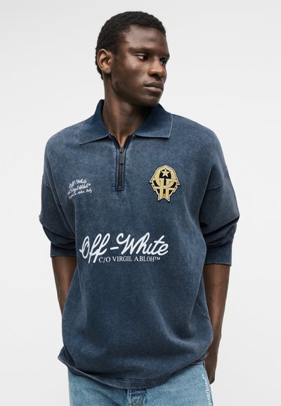 Νεαρός άνδρας που φοράει μπλε ναυτικό φούτερ Off-White με φερμουάρ μέχρι τον λαιμό, με λογότυπα και γυρισμένα μανίκια, κοιτάζει προς τα δεξιά του σε απλό φόντο.