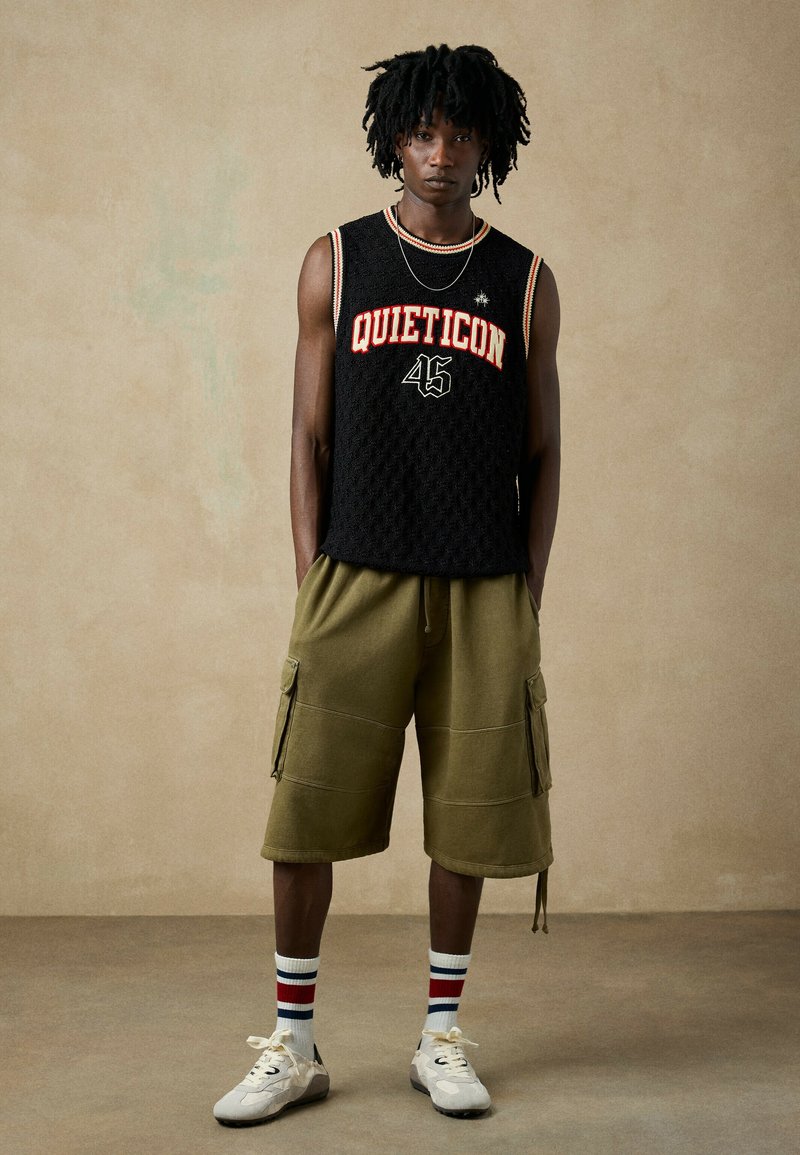 Jeune homme avec dreadlocks portant un haut noir sans manches en tricot avec "QUIETICON 45," shorts cargo olive, chaussettes rayées et baskets blanches, debout devant un fond uni.