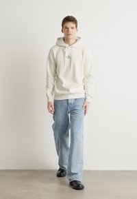 Sudadera crema con cordones y logo en el pecho, combinada con jeans de mezclilla azul claro y zapatos negros tipo slip-on. Fondo liso.