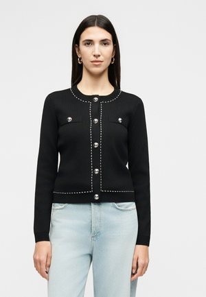 Femme portant un cardigan noir avec des boutons argentés et des surpiqûres blanches, associé à un jean bleu clair, debout devant un fond uni.