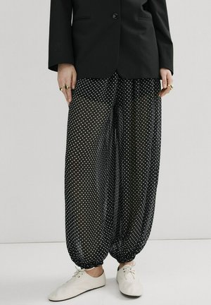 Dolna część ciała ubrana w czarną marynarkę, prześwitujące czarne spodnie w białe kropki, białe buty na sznurowadła oraz złote pierścionki na obu dłoniach.