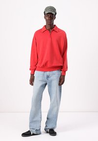 Felpa rossa con zip e colletto, abbinata a jeans azzurri chiari e sneakers nere. Modello indossa anche un cappellino verde. Texture morbide.