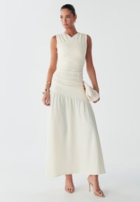Modèle portant une robe maxi sans manches, de couleur crème, se tient avec confiance, tenant une pochette texturée, avec des accessoires minimalistes et des cheveux lisses.