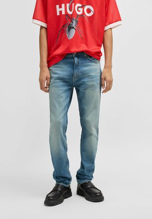 Blue denim jeans with a faded design, paired with a red graphic t-shirt featuring a black print. Black slip-on shoes complete the outfit.