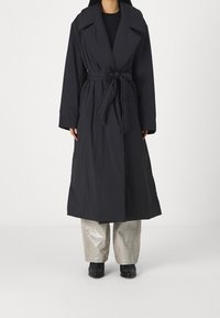 Abrigo largo y oscuro hecho de tela texturizada, con un amplio cuello y cinturón de lazo. El abrigo tiene un ajuste relajado con mangas anchas y un diseño hasta el tobillo.