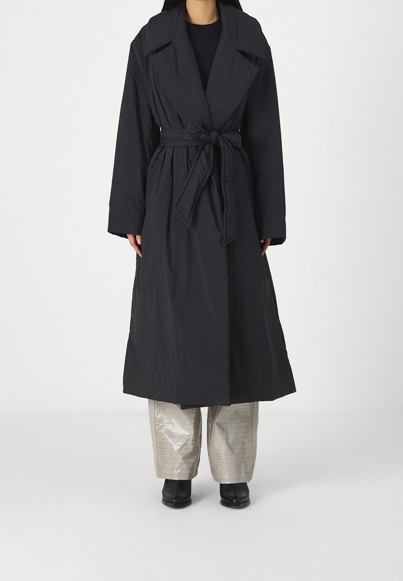 Abrigo largo y oscuro hecho de tela texturizada, con un amplio cuello y cinturón de lazo. El abrigo tiene un ajuste relajado con mangas anchas y un diseño hasta el tobillo.
