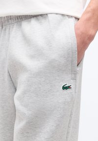 Ανοιχτό γκρι φόρμα με ελαστική μέση, ένα χέρι στην πλαϊνή τσέπη και μικρό πράσινο λογότυπο κροκοδείλου κοντά στην τσέπη.