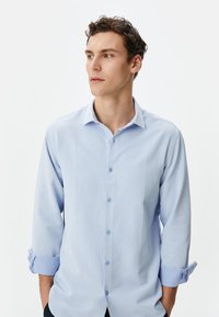 Camisa de manga larga azul claro con un patrón texturizado, cuello abotonado y mangas enrolladas. Tejido suave con un ligero brillo.