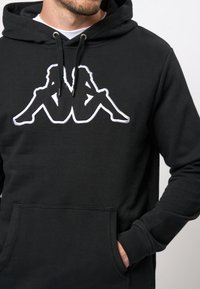 Zwarte hoodie van een katoenmix, met een groot wit geborduurd ontwerp van twee zittende figuren. Inclusief een voorzak en een verstelbare capuchon.