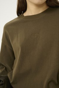 Nærbillede af en person iført en olivengrøn crewneck-trøje med et lille broderet logo centreret på brystet.