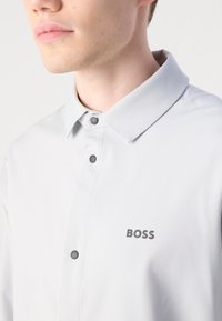 Jeune homme portant une chemise gris clair à boutons avec col, avec le mot "BOSS" imprimé sur la poitrine.