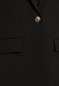 Blazer nero in tessuto morbido, con rever strutturati e chiusura a un bottone. Include due tasche frontali con patta.