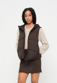 Gilet imbottito marrone con cappuccio, dotato di zip frontale e tasche laterali, indossato sopra un maglione lavorato a maglia beige chiaro e una gonna marrone.
