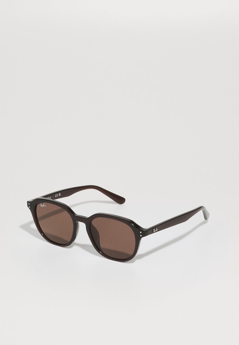 Lunettes de soleil noires en plastique avec des verres marron, présentant une forme géométrique arrondie, une légère épaisseur et des accents de logo argenté sur les branches.