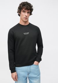 Sweat-shirt noir à col rond et manches raglan, avec texte blanc "CRAFTED GOODS PIER ONE DENIM." Porté avec un jean bleu clair.