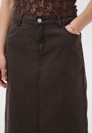 Personne portant une jupe en jean brun foncé avec un bouton devant, un haut en dentelle brun et un bracelet doré, main dans la poche, vue de la taille vers le bas.