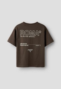 Brūna kokvilnas T-krekls ar baltu tekstu: "ROMA*" un "VIA DEI FORI IMPERIALI", ar kasti ražošanas informācijai aizmugurē.