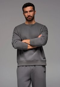 Sudadera de algodón gris con puños y dobladillo acanalados, con cuello redondo y un pequeño logo en el lado inferior. Combinada con pantalones a juego.