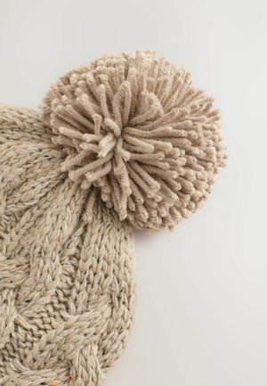 Beige Strickmütze mit strukturiertem Muster, ausgestattet mit einem großen, flauschigen Pompon aus weichem Garn auf der Oberseite. Keine sichtbaren Etiketten oder Akzente.
