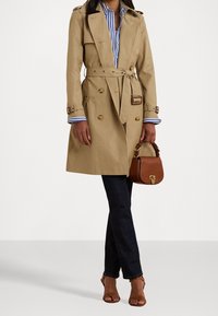 Beżowy płaszcz trencz z dwurzędowym zapięciem, paskiem w talii i pagonami na ramionach. Połączony z ciemnymi dżinsami i brązową torebką. Widoczna pasiasta koszula.