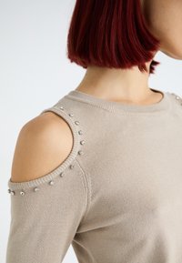 Maglione beige lavorato a maglia con spalle scoperte, adornato da strass lungo le cuciture delle spalle e del collo. Texture morbida, design aderente.