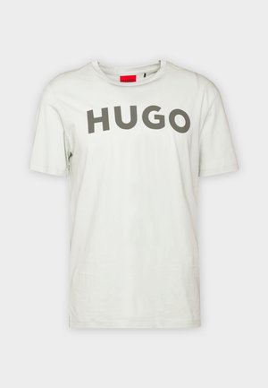 T-shirt bianca a maniche corte con grande scritta grigia "HUGO" sul petto e etichetta interna rossa sul colletto.