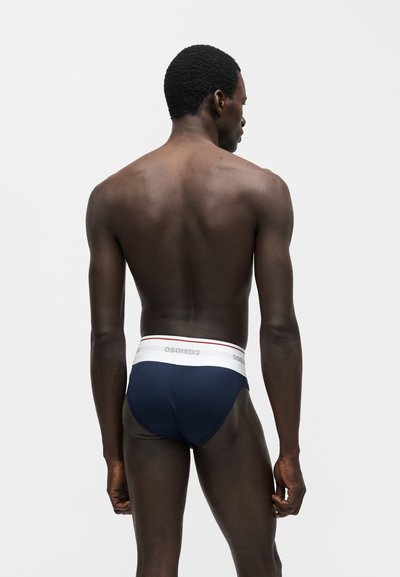 Άνδρας που στέκεται με την πλάτη, φοράει σκούρα μπλε εσώρουχα με λευκή ζώνη με την ένδειξη "DSQUARED2" σε απλό φωτεινό φόντο.