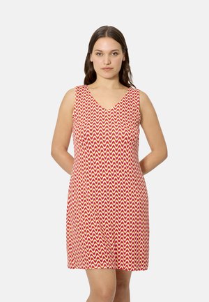 Mujer con vestido sin mangas hasta la rodilla con patrón de zigzag en rojo, naranja y blanco, de pie frente a un fondo liso.