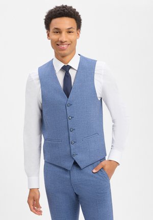 Lächelnder Mann, der eine hellblaue Weste und passende Hose, ein weißes Hemd und eine dunkelblaue Krawatte trägt, mit einer Hand in der Tasche stehend.