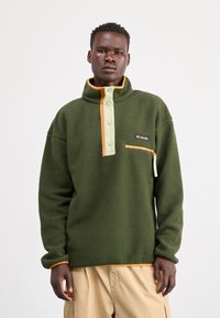 Mand iført grøn fleecepullover med orange kant og lynlåslomme, stående foran en ensfarvet hvid baggrund.