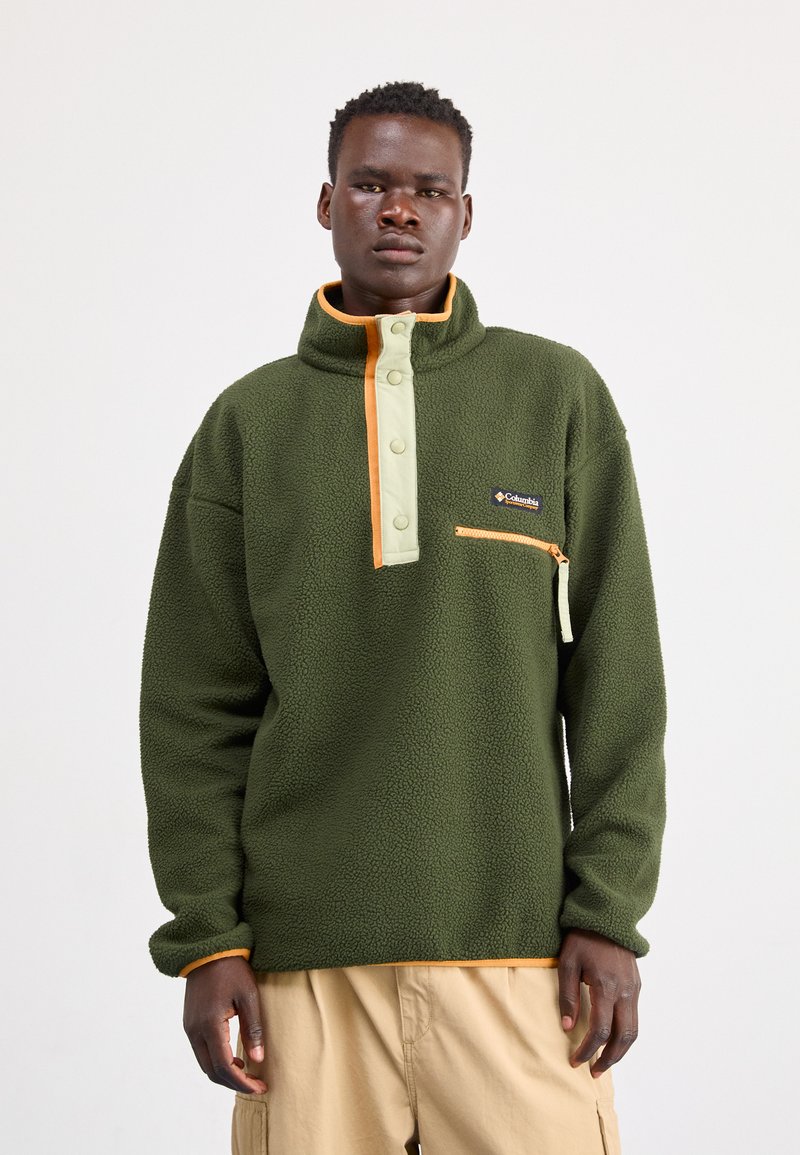 Mand iført grøn fleecepullover med orange kant og lynlåslomme, stående foran en ensfarvet hvid baggrund.