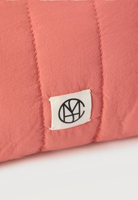 Εγγύς πλάνο υφάσματος σε κοραλί ροζ απόχρωση με ραμμένα σχέδια και μια μικρή λευκή ορθογώνια ετικέτα που φέρει ένα μαύρο αφηρημένο κυκλικό λογότυπο.