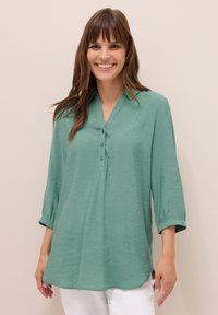 Tunique légère turquoise avec un décolleté en V, des manches trois-quarts, une patte de boutonnage à trois boutons et un ourlet légèrement arrondi. Texture douce.