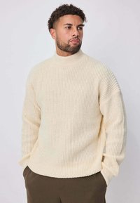 Pull tricoté couleur crème avec col montant, texture côtelée, épaules tombantes et coupe décontractée. Porté sur un pantalon vert olive.