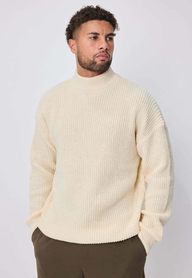 Pull tricoté couleur crème avec col montant, texture côtelée, épaules tombantes et coupe décontractée. Porté sur un pantalon vert olive.