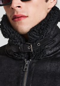 Primo piano di una persona che indossa una giacca in shearling nera con zip frontale e colletto con fibbia, mostrando una parte del viso e del collo.