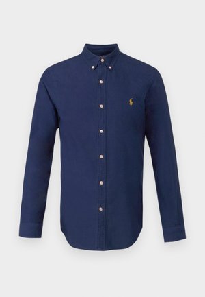 Chemise à manches longues bleu marine en coton, avec un col boutonné et un petit logo doré sur la poitrine. Coupe classique avec patte de boutonnage.