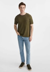 Oliwkowa krótko rękawowa koszulka z dzianiny z kieszonką na piersi, w zestawie z jasnoniebieskimi prostymi dżinsami i ciemnozielonymi trampkami.