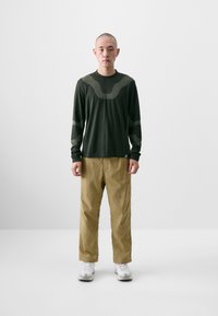 Long-sleeve, dark green shirt with textured design and lighter accents, paired with beige pants and white sneakers, standing on a neutral background.
