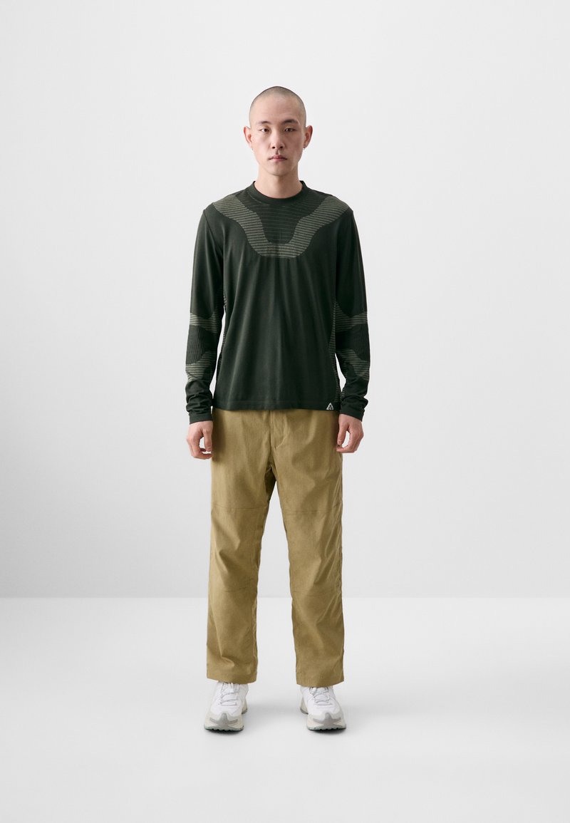 Langarm-Hemd in Dunkelgrün mit strukturiertem Design und helleren Akzenten, kombiniert mit beigen Hosen und weißen Sneakers, vor neutralem Hintergrund stehend.
