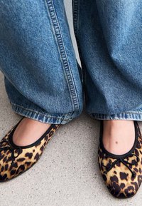 Ballet flats in a leopard print pattern with a black trim and bow detail, paired with blue wide-leg jeans against a light surface.