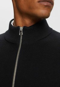Homme portant un pull zippé côtelé noir avec un col montant, fermeture éclair remontée jusqu’au cou, montrant une partie de son bas du visage et de son cou.