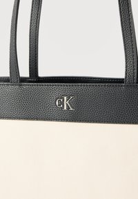 Μαύρη και λευκή τσάντα tote με υφασμένους μαύρους λαβές και περιμετρικό τελείωμα, με ασημένιο λογότυπο "cK" στην πάνω μπροστινή πλευρά.